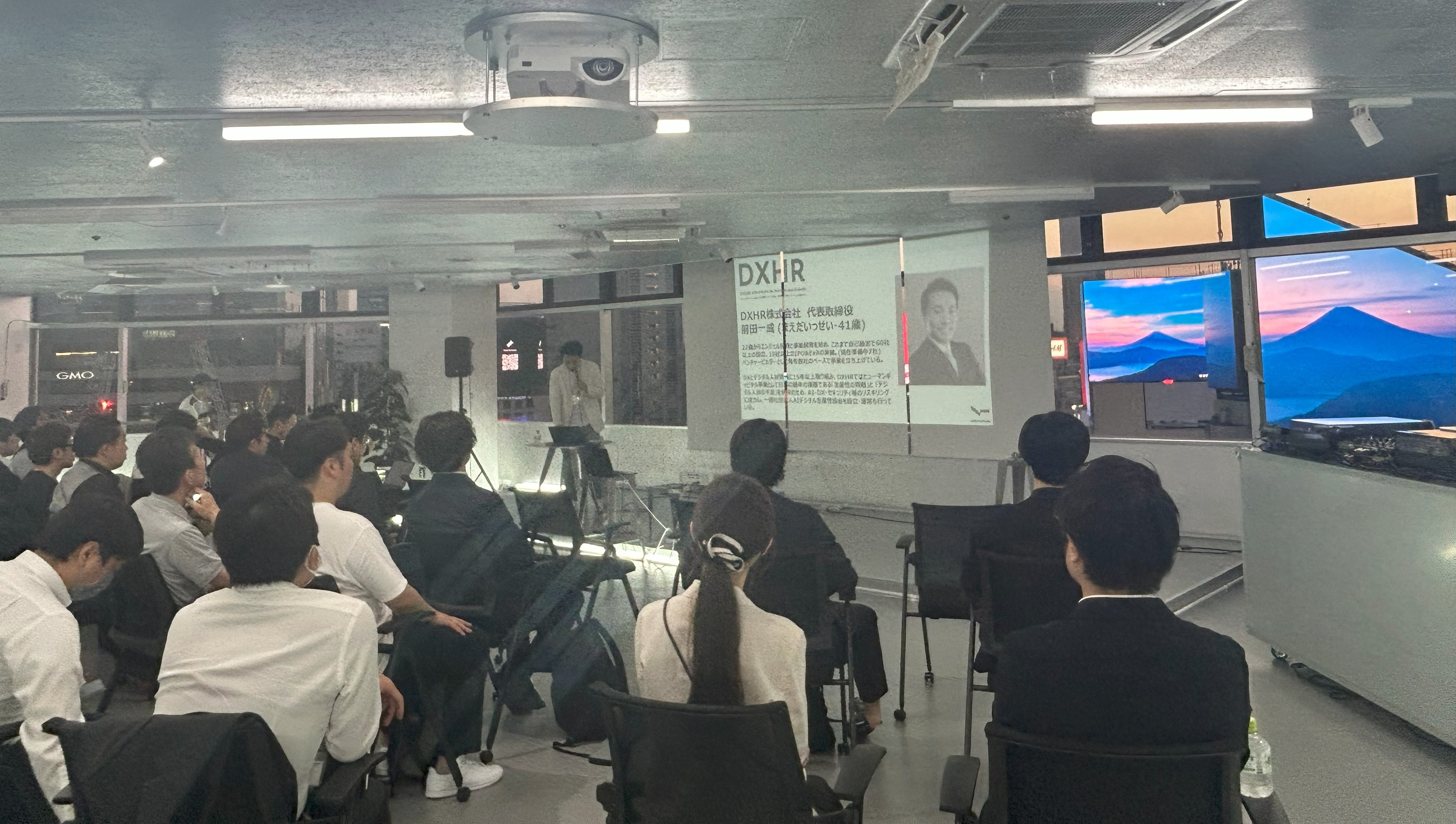 登壇者がスクリーンの横で発表をしている場面。スクリーンには「DX×HR」のタイトルと代表取締役のプロフィールが映し出されている。