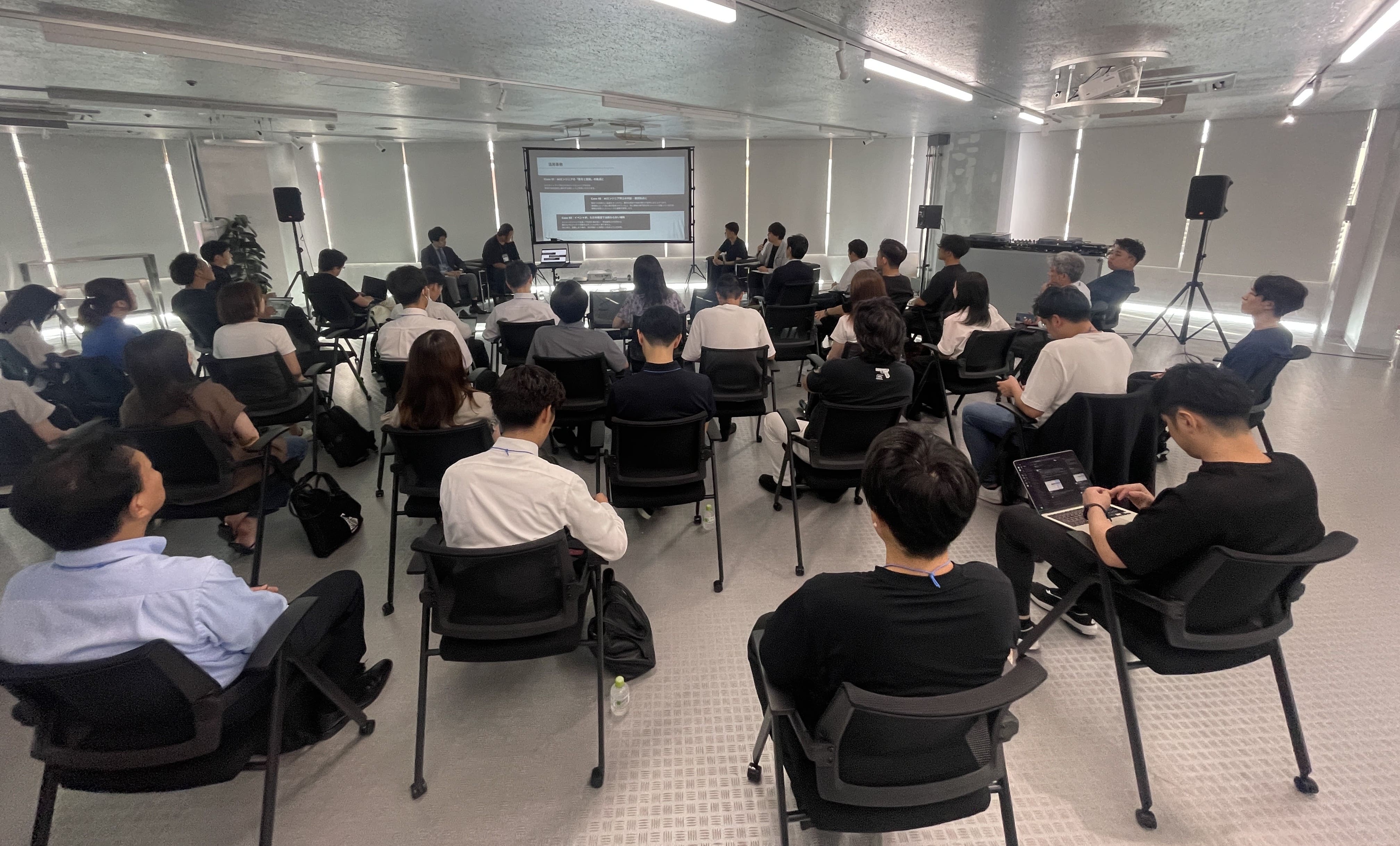 会議室で開かれているパネルディスカッションの様子。スクリーンに資料が投影され、前方に登壇者が並んで座っている。聴衆は20〜30名ほどで、椅子に座って発表を聞いている。